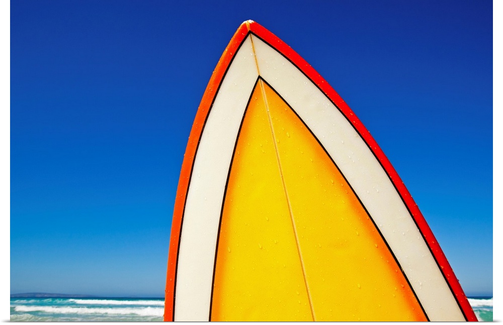 The very top of a surfboard is photographed with a view of the deep blue sky and ocean just behind it.
