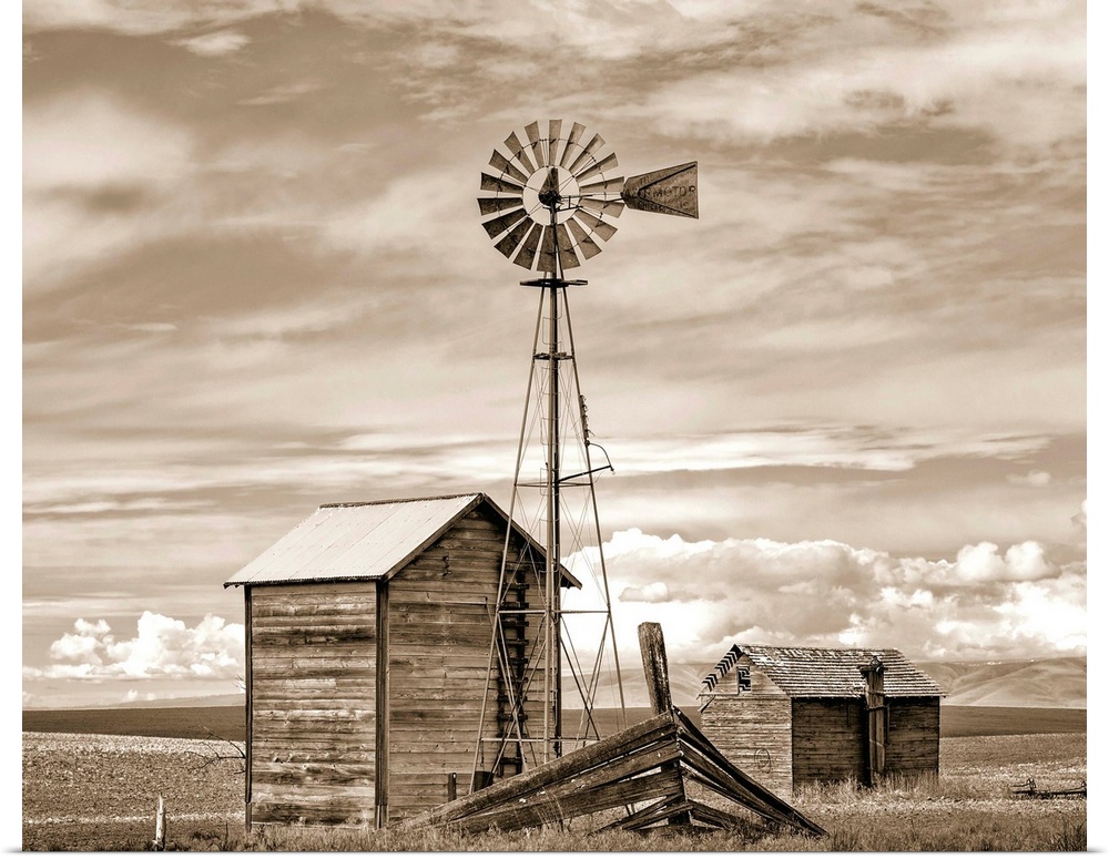 A sepia photograph of a windmill and shed in a field with layers of clouds overhead.