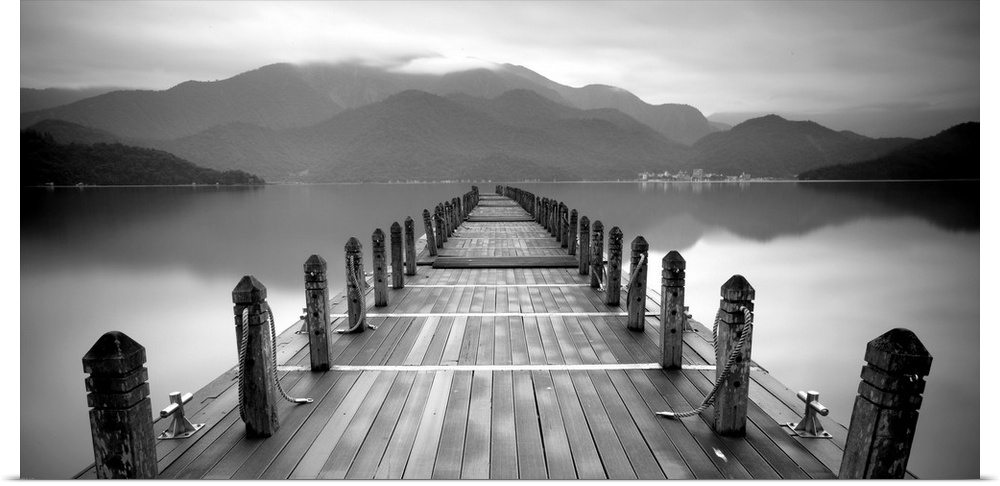 A black and white photograph of a long pier at a lake with large mountains in the distance.