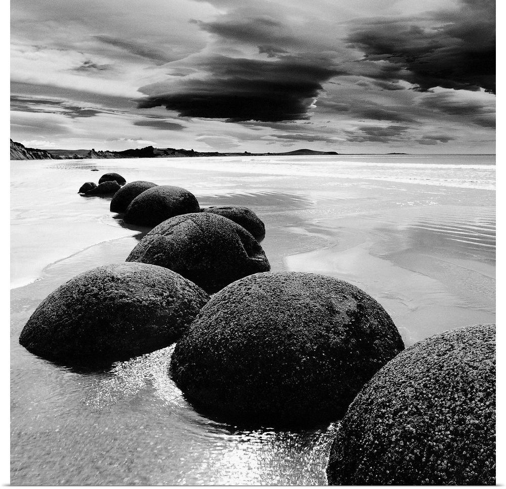 Black and white photograph of a row of smooth rocks on a beach with a dramatic sky full of clouds.