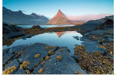 The rocky peak of Volanstinden lighted up by the midnight sun is reflected in blue sea