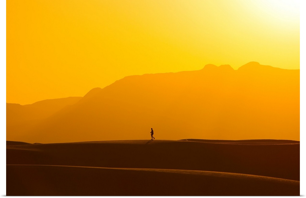 A lone man walks on top of sand dunes during sunset.