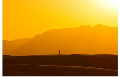 A lone man walks on top of sand dunes during sunset