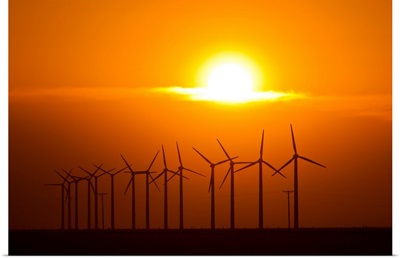 The sun sets behind a row of spinning windmills or wind turbines