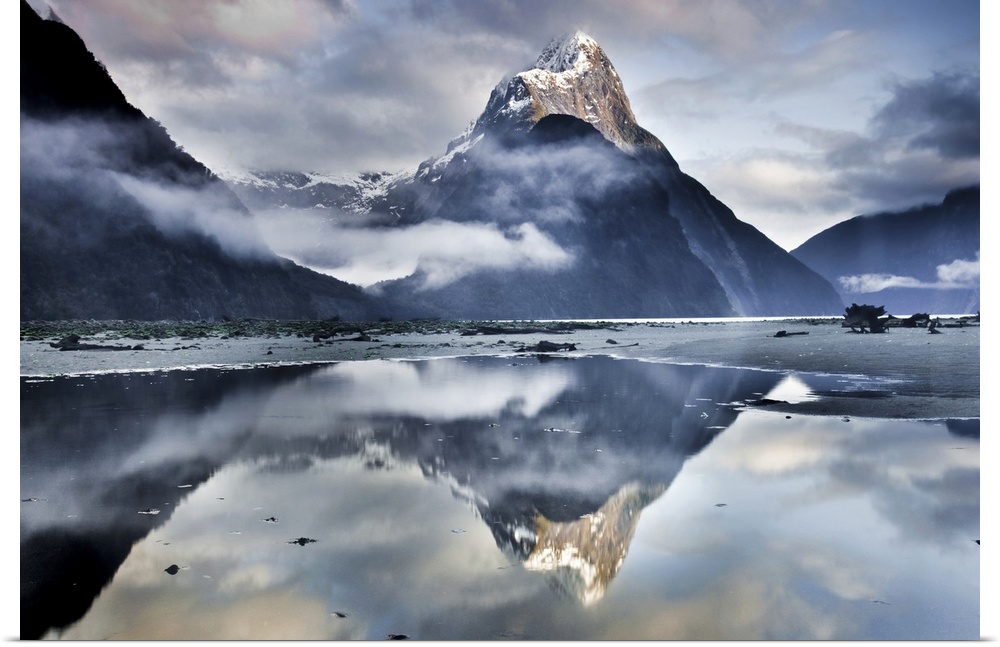 A landscape photograph of a snow covered mountain peak and misty clouds reflecting in shallow beach waters.