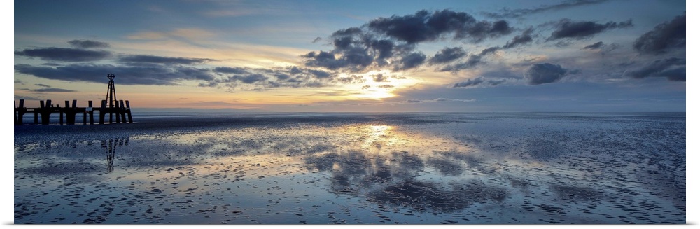 A panoramic image of a tranquil golden blue dawn with fluffy clouds over reflecting wet sand with a harbour installation.