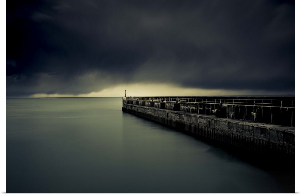 A photograph of a pier jetting out over water under a sky filled with a blanket of dark clouds.