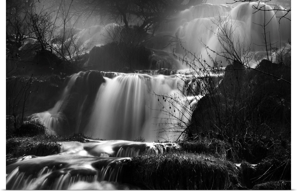 This black and white photograph is taken of several small waterfalls that are surrounded by bare trees and foliage.