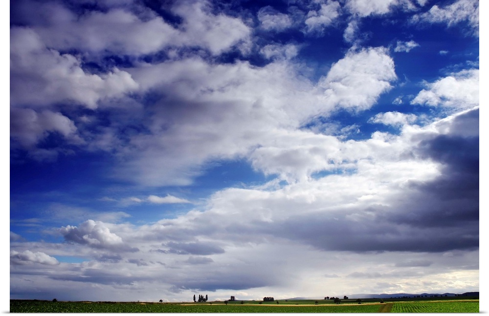 Large horizontal photograph of many fluffy white clouds in a vibrant blue sky, over a green farm landscape.