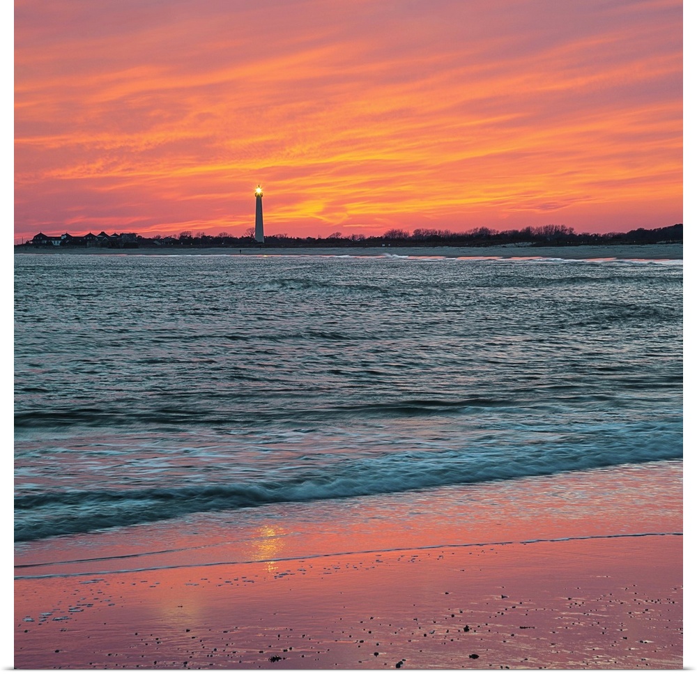 Vivid orange sunset sky over the ocean at low tide, with Cape May lighthouse shining in the distance, Cape May, New Jersey.