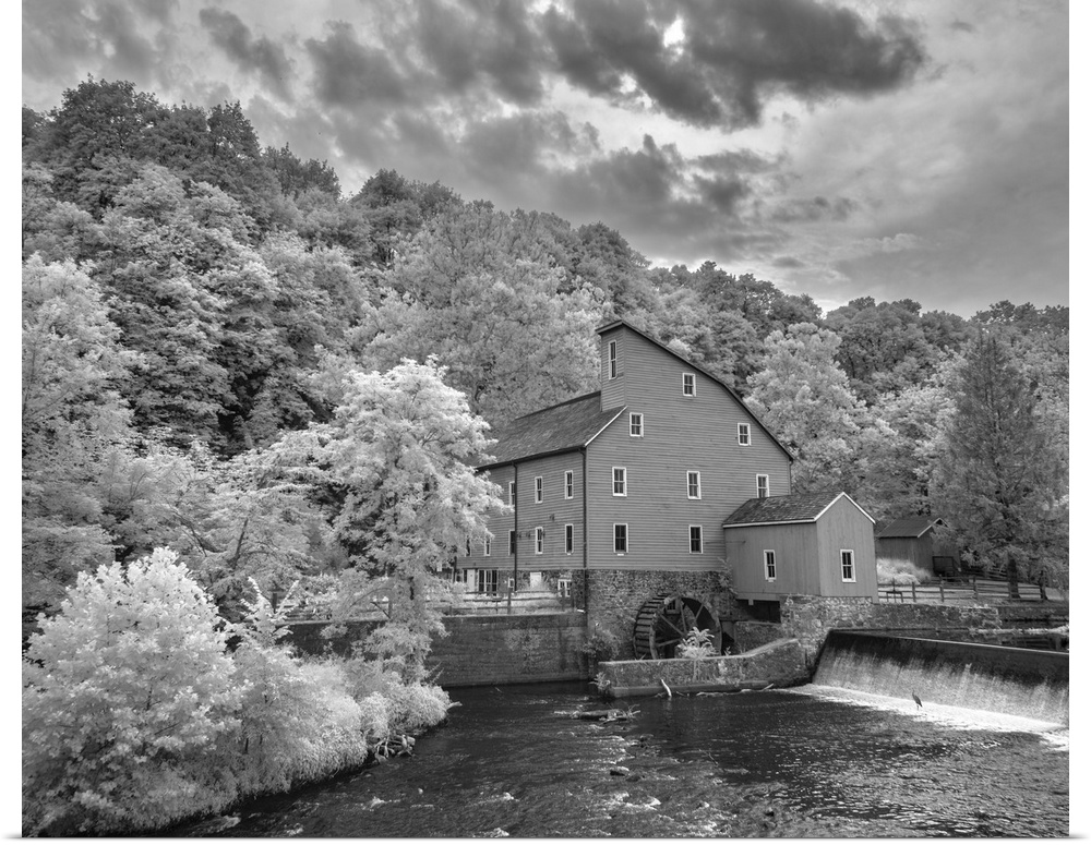 In this black and white infrared light photograph, dramatic and brightly lit clouds rest over a beautiful old mill, nestle...