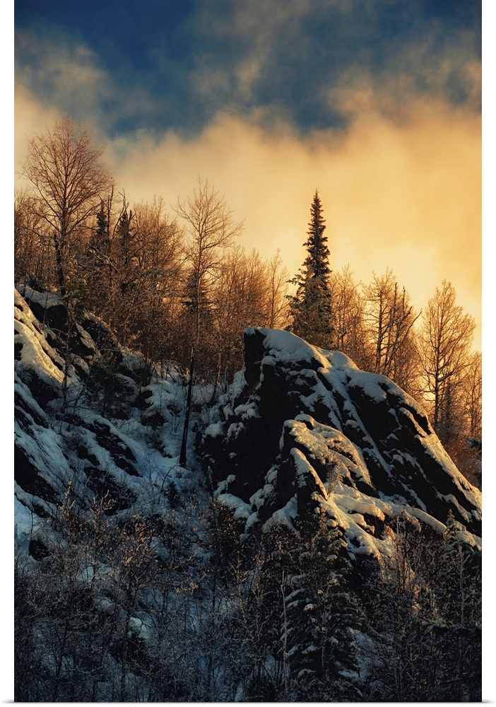 Clouds lit up with fading sunlight over a forest near snowy rocks on a mountainside.