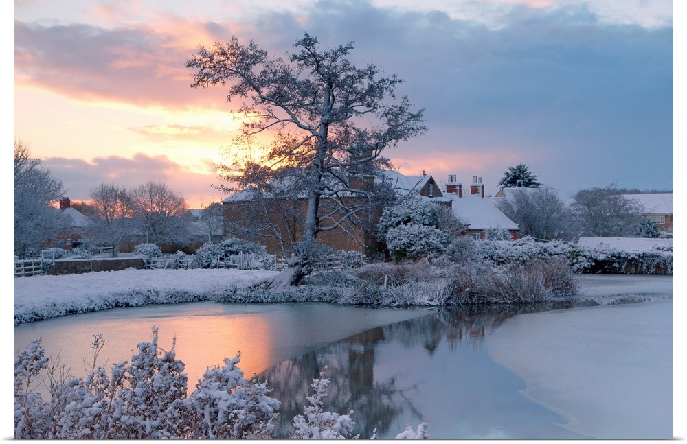 A golden dawn over a village pond and snowy fields to the village roofs beyond with trees in snow, Nottinghamshire, UK.