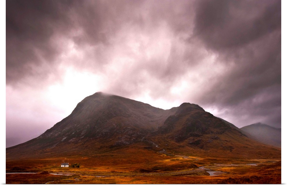 A majestic Scottish mountain in Glencoe with dramatic storm clouds and rich autumn colours with a small white cottage dwar...