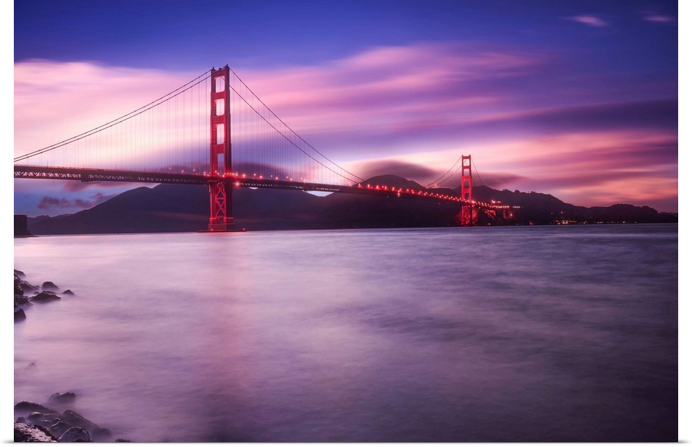 Landscape photograph of a purple and pink sunset over the Golden Gate Bridge in San Francisco with calm waters below.