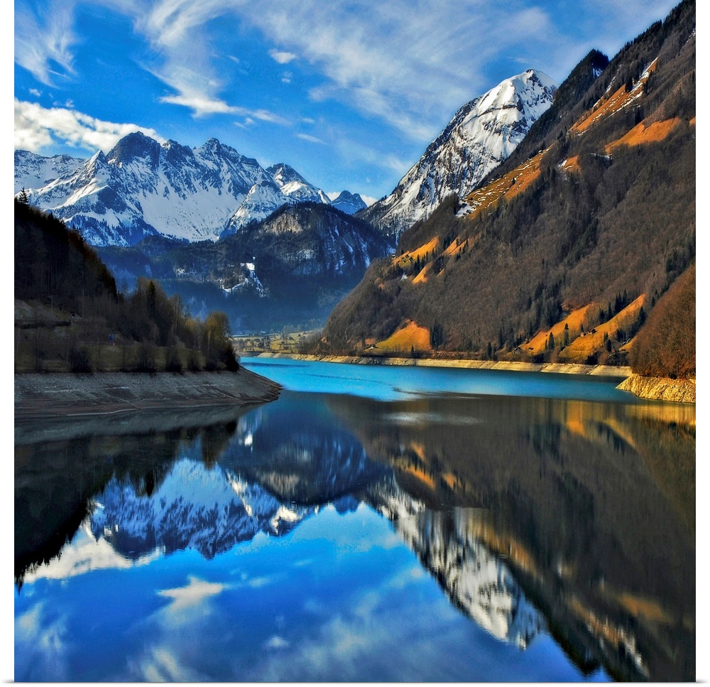 Square photograph on a big wall hanging of snow covered mountains beneath a blue sky, reflecting in still waters in the fo...