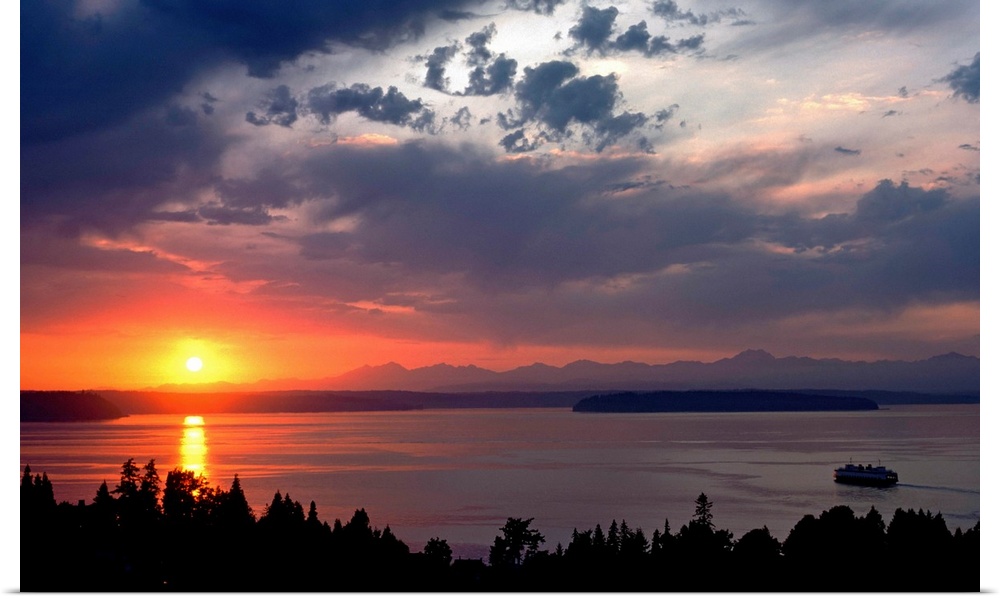 Photograph looking out over a body of water as the sun sets behind a distant mountain range creating a firey sunset.