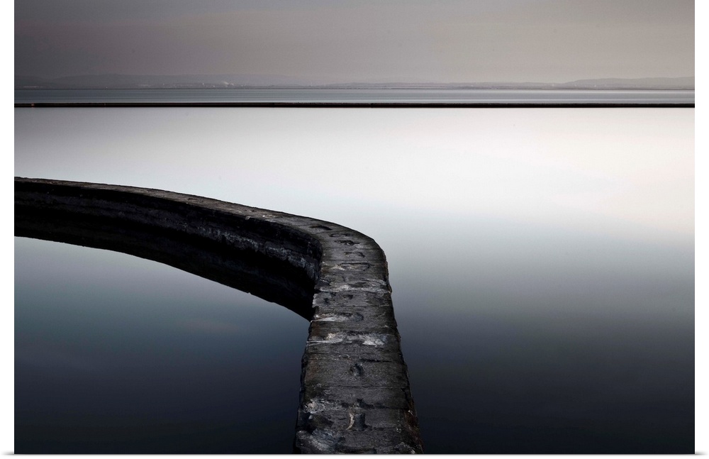 A monochrome zen-like minimalist image of mirror calm water in a pool with a curving wall and a distant horizon.