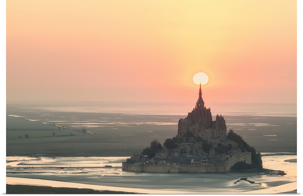 The sun appearing to rest on the spire at the top of Mont Saint Michel in France at sunset.