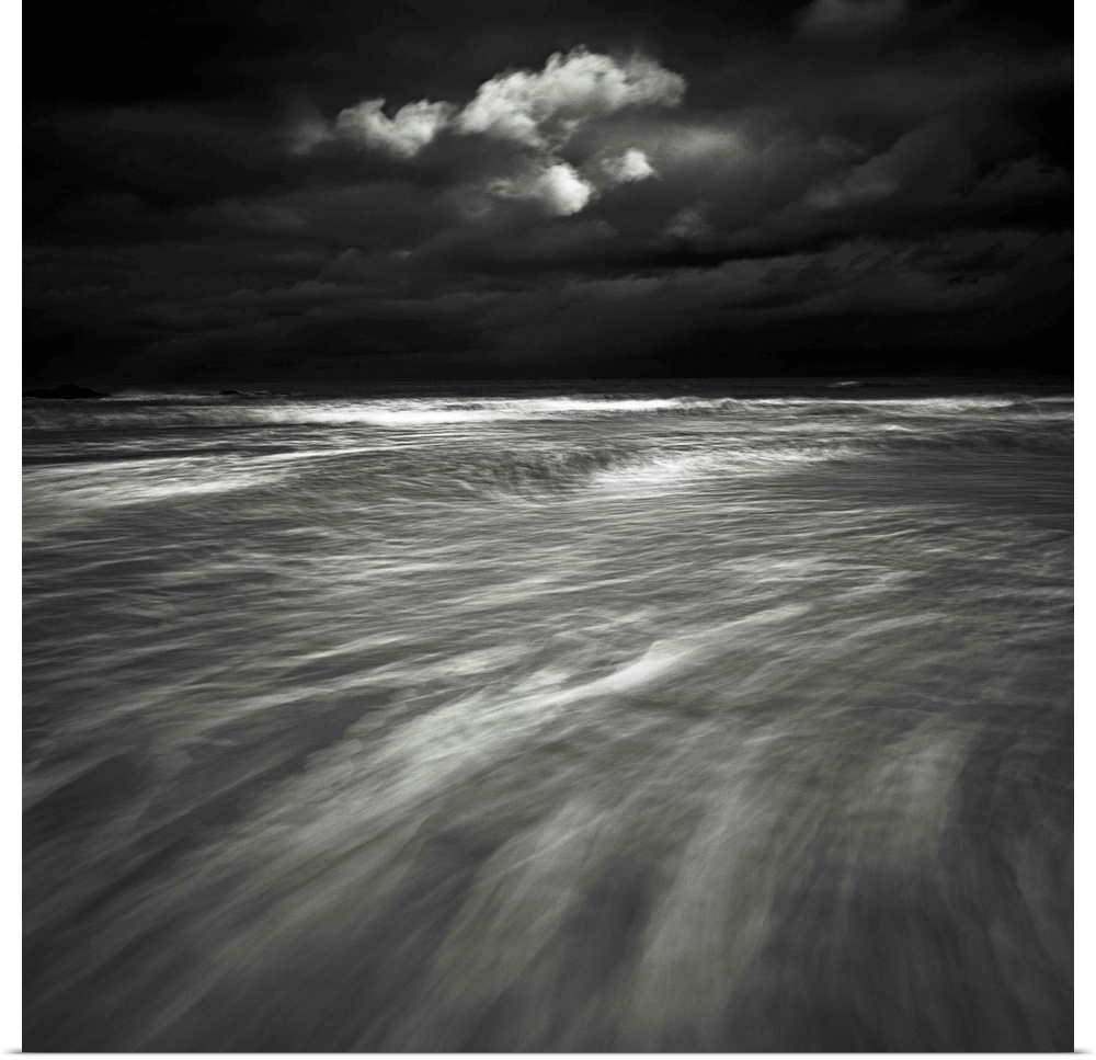 A monochrome black and white sepia toned dramatic image of swooshing wavws and a lone cloud in a dark stormy sky over a se...