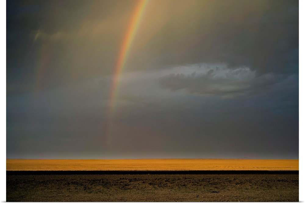 Landscape photograph of an open landscape with golden fields, a contrasting dark road, and a rainbow in the gray and blue ...