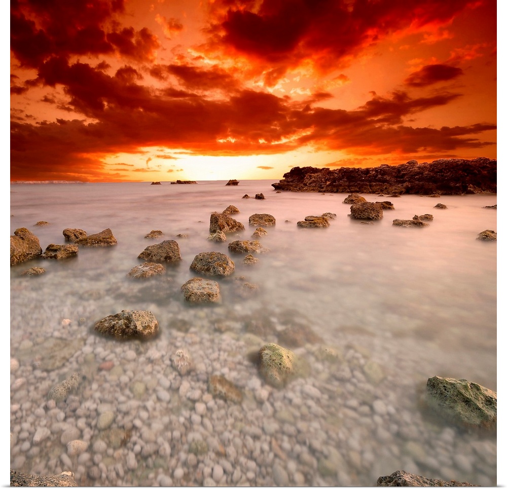 Square, fine art photograph of a shallow, large body of water full of rocks, beneath a deep orange and red sunset.