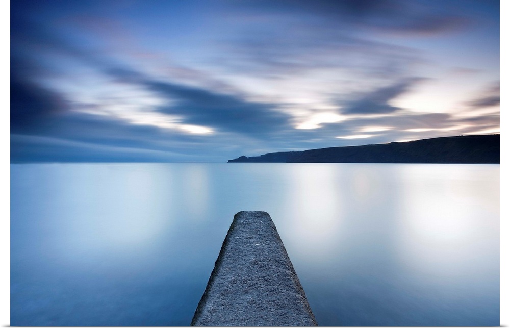 Tranquil calm blue seascape with symetrical jetty into a flat sea with floating clouds.