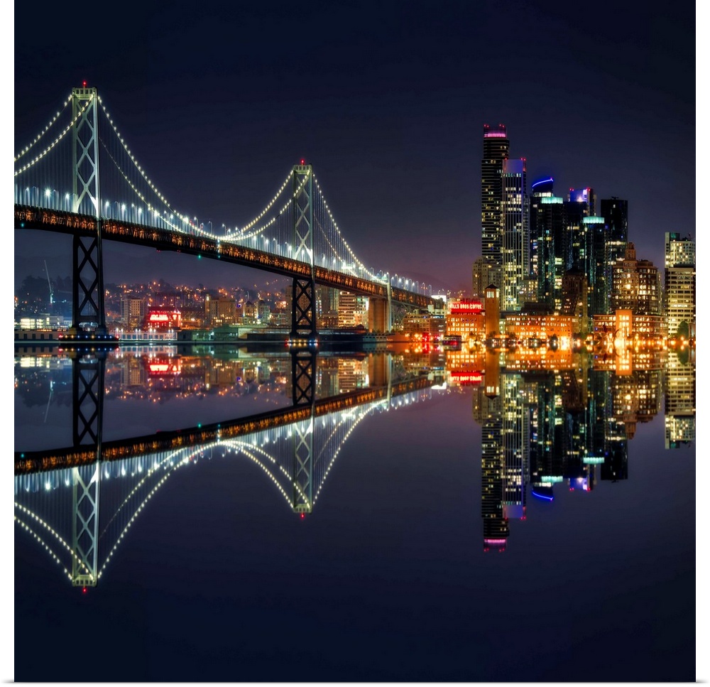 Square photograph of the Bay Bridge and downtown San Francisco lit up at night and reflecting onto the motionless water.