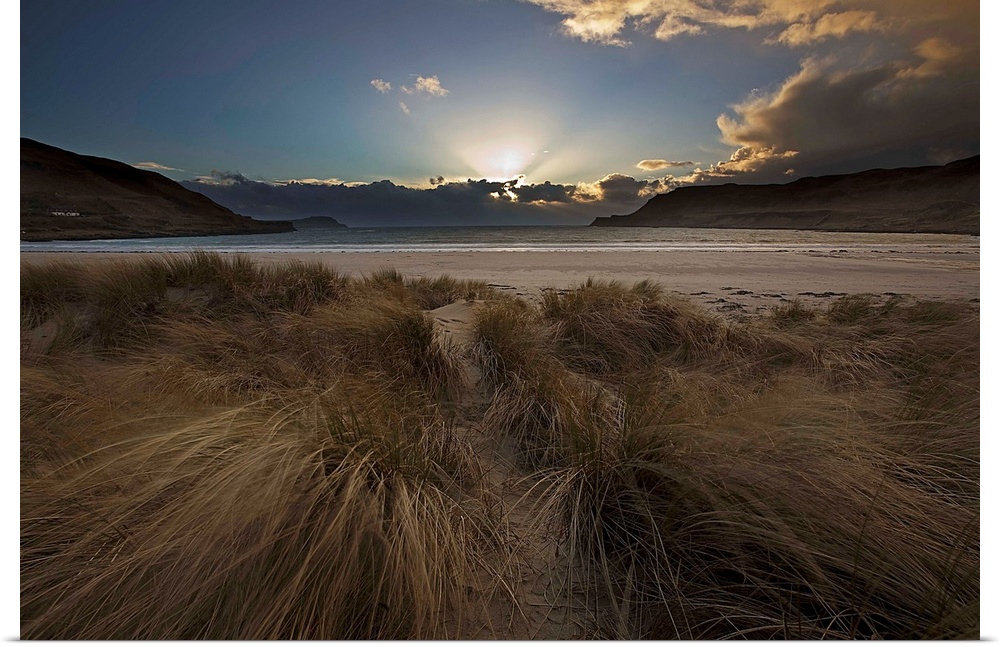 A dramatic seascape sunrise with crepuscular God's Rays from behind clouds with a blue sky and wind blown marram grass on ...