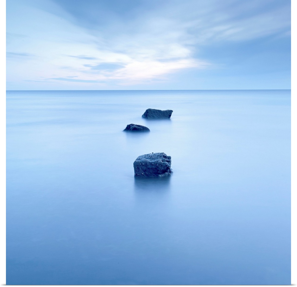 Large, square photograph of three rocks rising up from calm waters, beneath a partly cloudy sky.