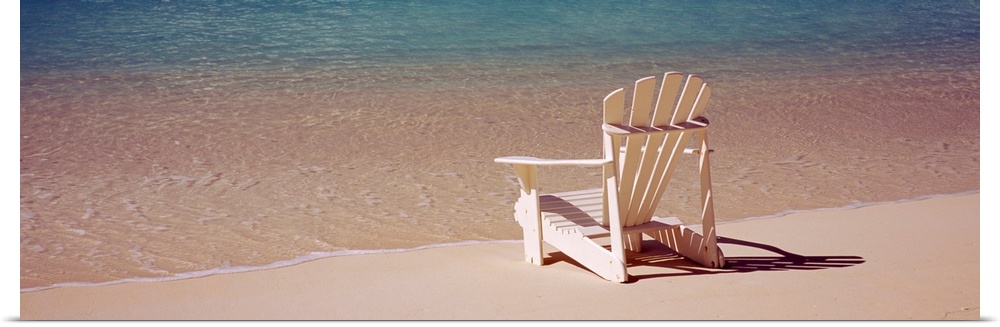 A wooden chair designed for relaxing reclining sits on the edge of the shore as waves wash over the sand around it. This p...