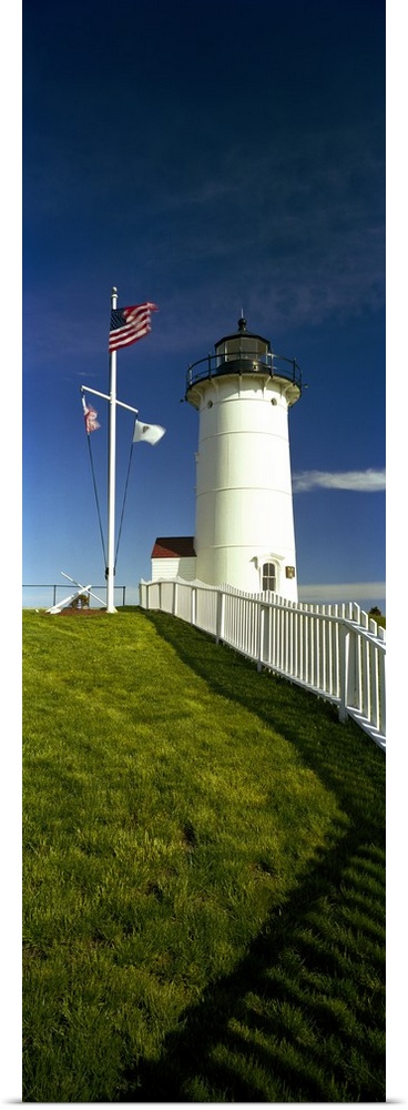 Giant vertical photograph looking up a grassy hill along a fence toward Nobska Lighthouse next to an American flag, agains...