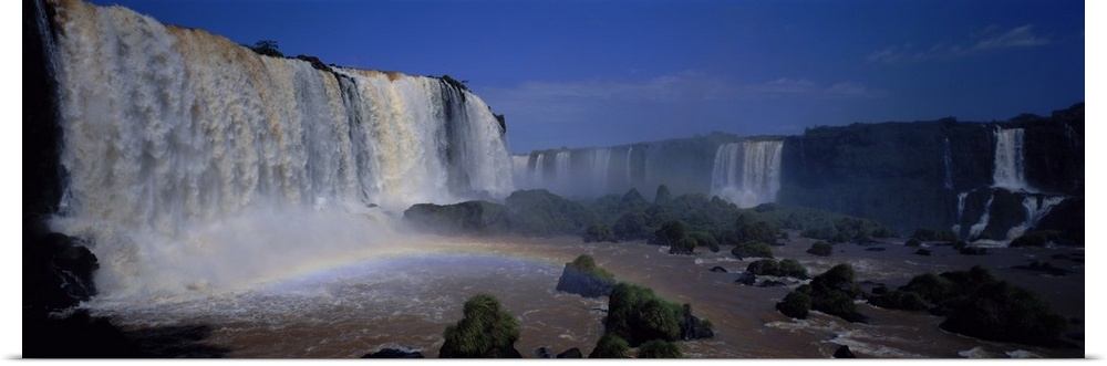 This is a panoramic photograph of enormous South American waterfalls and a rainbow forming under the cascades.