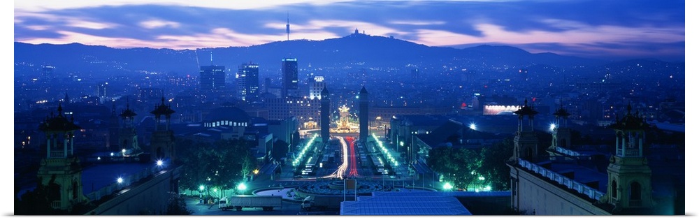 Giant, high angle photograph of Barcelona, lit at night, mountains in the background beneath a mostly cloudy sky, in Spain.