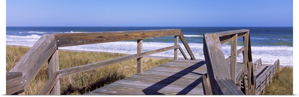 Panoramic picture taken of a small walking bridge headed down toward the beach with dunes on either side and the ocean str...