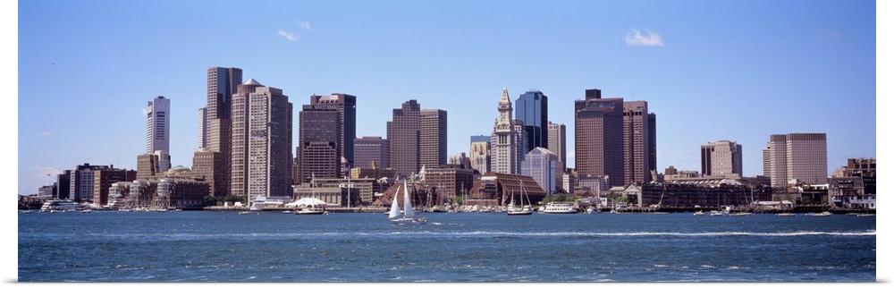 The Boston skyline is photographed from across the water on a beautiful sunny day with sail boats gliding in the water.