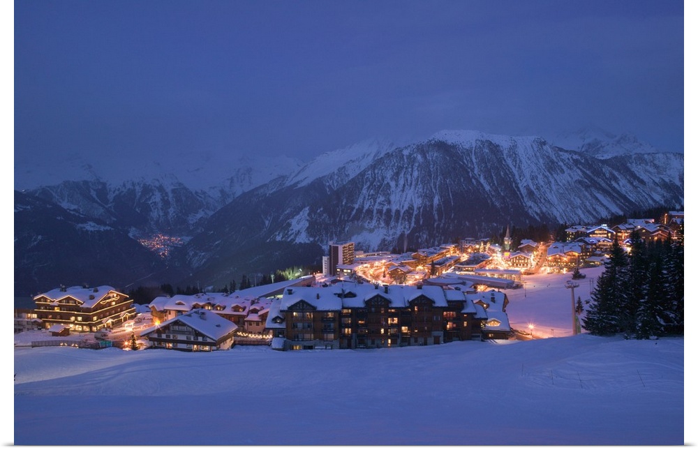 Photograph of small town nestled at the bottom of snow covered mountain at night.