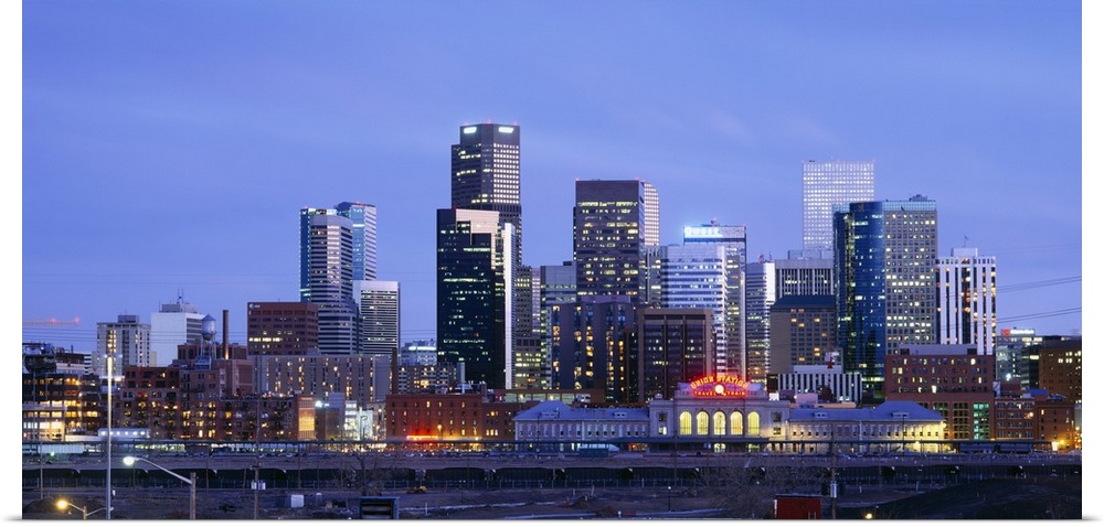 A large panoramic piece of a photograph of the Denver skyline with the buildings lit up during sun down.