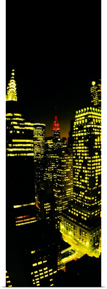 Vertical panoramic of lit up office buildings in Manhattan with the Empire State building peeking up in the distance in red.