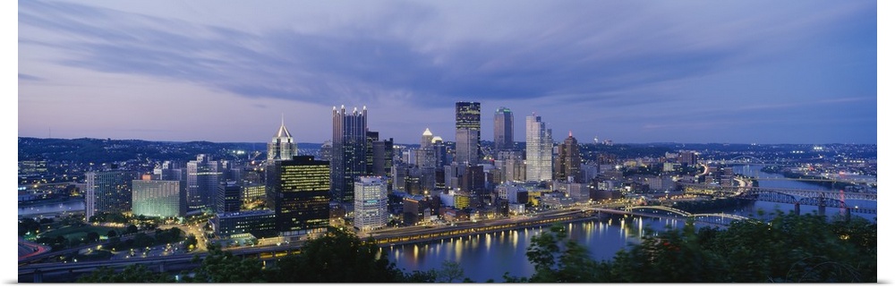 A beautiful photograph of the Pittsburgh skyline lit up at dusk. The river and trees can be seen in the foreground.