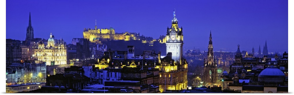 Panoramic photograph taken of buildings in Scotland lit up at night with a castle shown in the background.