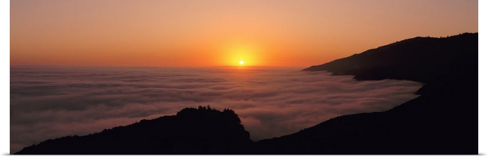 A large panoramic piece of a sunset with a fog mist over the water and mountains lining the right side of the picture.