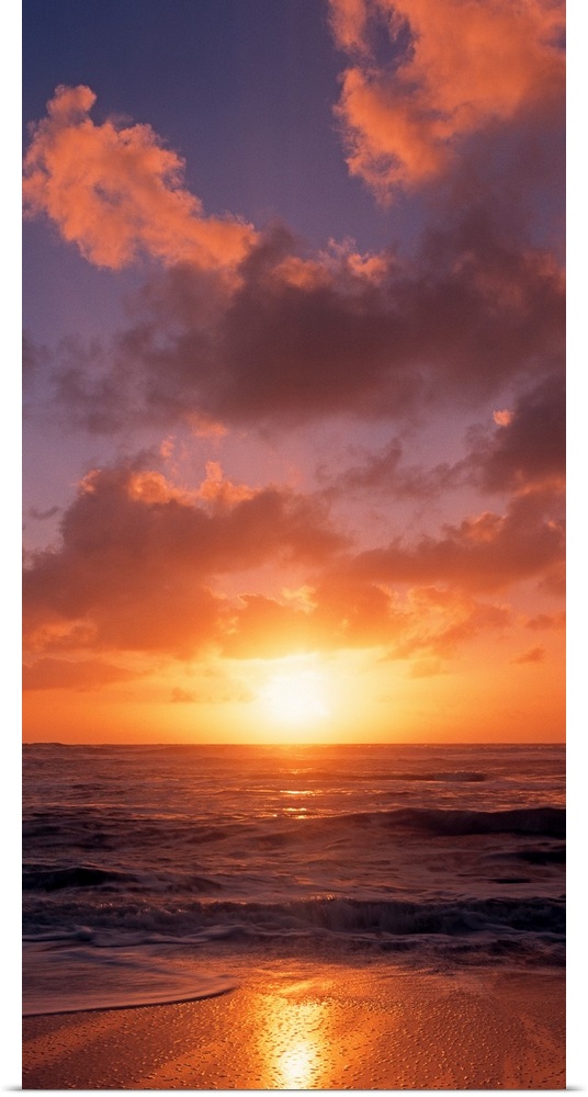 This vertical photograph shows the sun setting at the beach as waves wash against the shore.