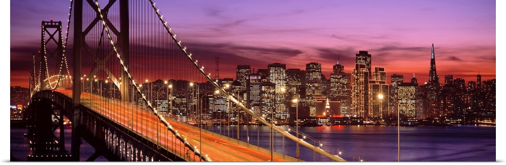 Panoramic photograph of the San Francisco Bay Bridge and the brightly lit city skyline at dusk.