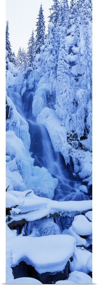 A tall panoramic piece of a frozen waterfall that is surrounded by trees and rocks that are covered with snow.