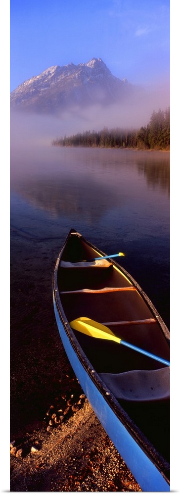 Tall and narrow canvas photo of a canoe half way in a lake with fog in the distance in front of a mountain.