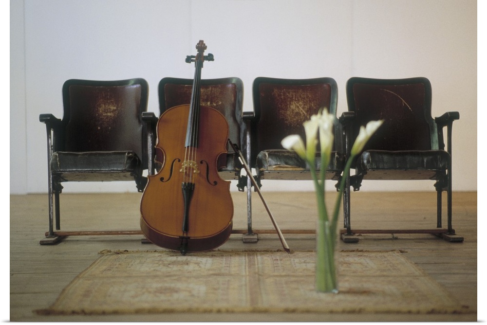 A cello stands on the floor and leans against a row of four seats. A vase of calla lilies sits on an area rug in front of ...