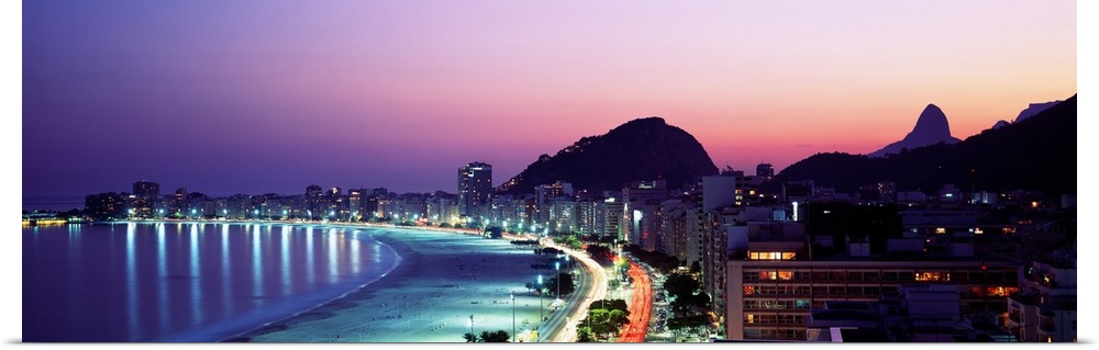 Panoramic, giant photograph of buildings lit along Copacabana Beach at night, mountains in the background against a vivid ...