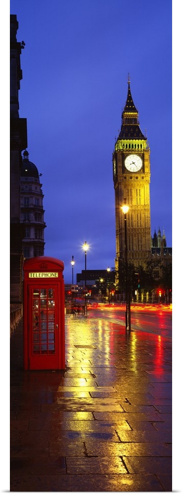 A long vertical photograph of Big Ben at night with an old fashioned telephone booth to the left of it on a stone sidewalk.