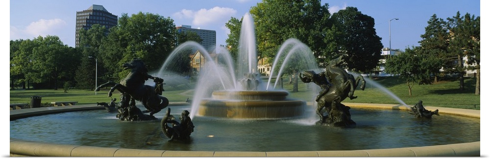 A large fountain with various statues in it is photographed in panoramic view with a park shown in the background.